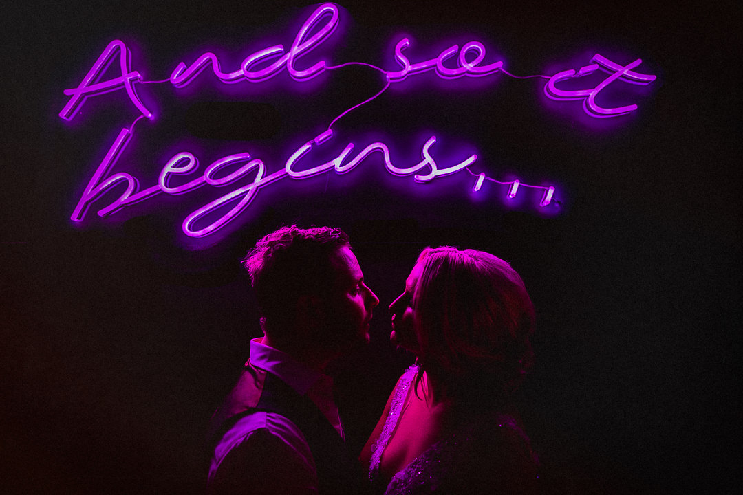 a couple embrace and are lit by a neon sign saying and so it begins
