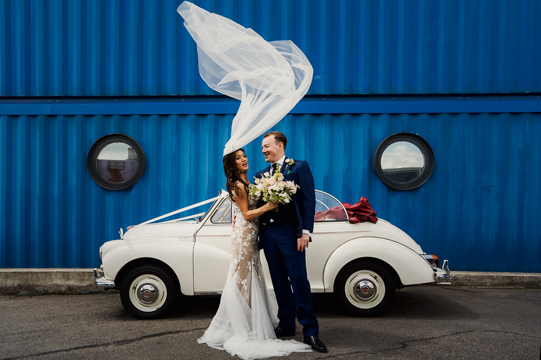 a brides veil blows upwards before a portrait with her husband and wedding car against a blue wall at trinity buoy wharf