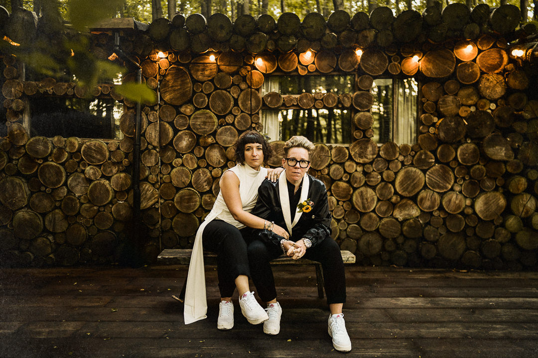 A same sex couple sit for a wedding portrait outside a log cabin at the dreys wedding venue